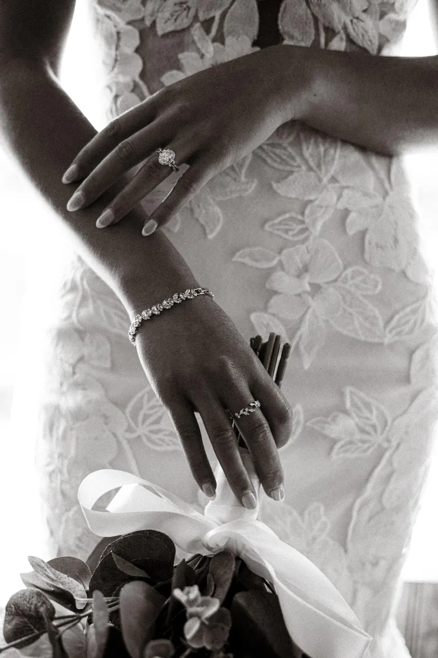 Black and white detail of a bride's hands, bracelet and lace dress above her bouquet